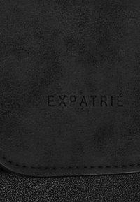 Materiale in pelle nera con superficie texturizzata, caratterizzato da un logo embossato "EXPATRIÉ." Patta liscia con cuciture visibili lungo i bordi.