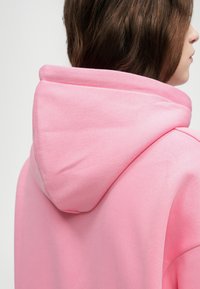 Rosa Kapuzenpullover mit weicher Textur und großer, runder Kapuze. Verfügt über ein glattes Design mit dezenten Nähten, vor einem neutralen Hintergrund.