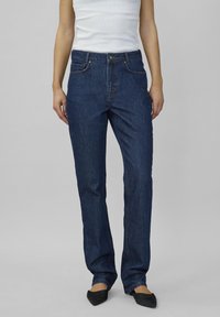 Žena v tmavomodrých rovno strihaných džínsoch, bielom bezrukávovom tope a čiernych špicatých balerínach stojí na pozadí v svetlo sivej farbe.