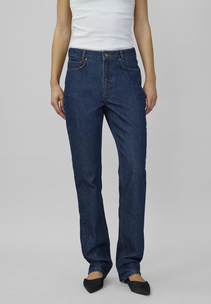 Žena v tmavomodrých rovno strihaných džínsoch, bielom bezrukávovom tope a čiernych špicatých balerínach stojí na pozadí v svetlo sivej farbe.