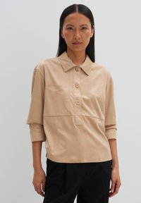 Lichtbeige katoenen shirt met een knoopsluiting aan de voorkant, een borstzak en driekwartmouwen. Zachte textuur en klassiek kraagontwerp.