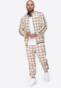 Beige kariertes Trainingsanzug mit kontrastierenden Streifen, elastischen Bündchen und Logo-Details, kombiniert mit weißen Turnschuhen.