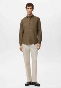 Camisa marrón abotonada con cuello, mangas largas y textura suave, combinada con pantalones beige claro y zapatos negros.