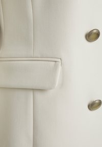 Crèmekleurige blazer van fijn geweven stof, met een zijzak en twee decoratieve metalen knopen aan de voorkant.