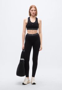 Σετ αθλητικό σουτιέν και κολάν σε μαύρο χρώμα με ελαστικές ταινίες με λογότυπο. Το μοντέλο κρατάει μια μαύρη τσάντα tote, φορώντας μπεζ αθλητικά παπούτσια. Ομαλή, εφαρμοστή υφή.