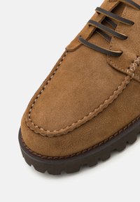 Loaferi din suede brun cu șireturi negre, cusături în contrast și talpă din cauciuc texturat. Vedere de aproape care subliniază materialul moale și forma.