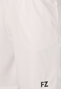 Närbild av vita sportshorts med svart "FZ"-logotyp nära fållen på höger ben.