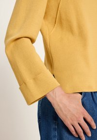 Person in einem gelben Langarmoberteil mit umgeschlagenen Ärmeln und blauen Jeans, die Hand ruht nahe der Hüfte.