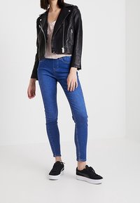 Schwarze Leder-Bikerjacke, hellrosa Oberteil, blaue Skinny-Jeans und schwarze Sneaker mit weißer Sohle. Das Model steht vor einem neutralen Hintergrund.