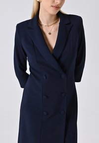 Donna che indossa un vestito blazer doppiopetto blu navy con collane in oro a catena e medaglioni, in posa su uno sfondo chiaro e semplice.