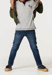 Grön bomberjacka med blommig foder, randig långärmad skjorta, blå denimjeans och svarta sneakers med gummisålar.