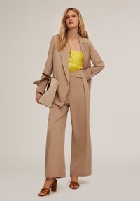 Beige op maat gemaakt pak met wijde broekspijpen en een aansluitende gele haltertop. Accessoires zijn een beige tas en bruine sleehakken.