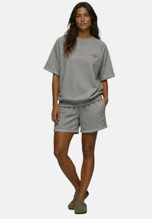 Femme debout portant un haut gris oversize à manches courtes et un short assorti avec de petits détails orange sur la fermeture éclair et des chaussures marron slip-on.