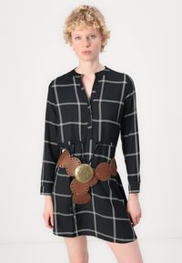 Rochie în carouri alb-negru, cu un decolteu cu nasturi, având un brâu din piele cu decupaje circulare și o cataramă metalică decorativă.