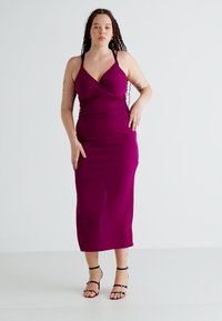 Prilagođena haljina srednje dužine od duboke magenta tkanine s preklopnim prednjim dijelom. Ima tanke naramenice i glatku, rastezljivu teksturu.
