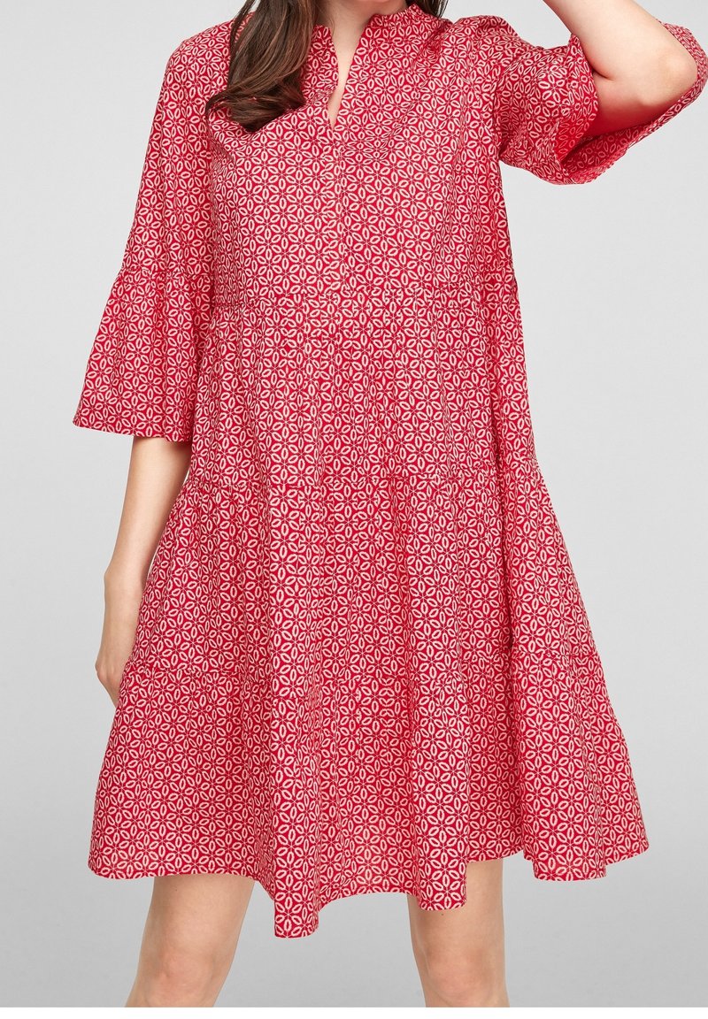 Robe rouge longueur genou avec motif floral géométrique blanc, manches cloches trois-quarts et encolure ronde avec une petite fente.