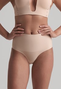 Donna che indossa biancheria intima beige alta in vita senza cuciture e un reggiseno coordinato, in piedi con le mani sui fianchi su uno sfondo neutro.