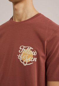 Bordeaux katoenen t-shirt met een ronde halslijn en korte mouwen. Een gedrukt ontwerp met cursieve tekst en een zon symbool staat op de linkerborst.