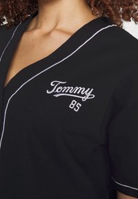 Maglietta nera a maniche corte con scollo a V, caratterizzata da una bordatura bianca lungo i bordi e con ricamo "Tommy 85" sul lato sinistro del petto.