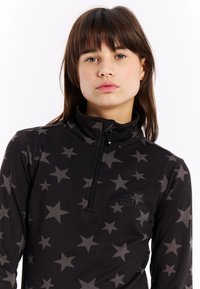 Haut zippé noir arborant un motif d'étoiles dans des nuances de gris variées. Fabriqué à partir d'un tissu lisse avec un col haut.