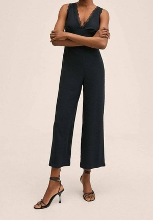 Musta ilmanhiha jumpsuit, jossa on pitsireunus, leveät lahkeet ja valmistettu sileästä kankaasta. Yhdistetty mustiin hihnallisiin korkeakorkoisiin sandaleihin.