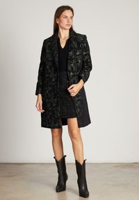 Mujer con un abrigo negro de patrón floral sobre una blusa negra de escote en V y una falda brillante, combinada con botas de cuero negras hasta la mitad de la pantorrilla, de pie en el interior.