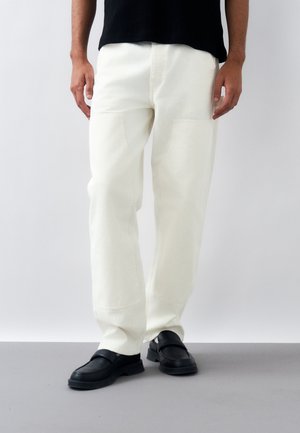 Άνδρας που φοράει φαρδιά λευκά παντελόνια με μεγάλες μπροστινές τσέπες και μαύρα παπούτσια τύπου σλίπ-ον, στέκεται μπροστά σε απλό φόντο.