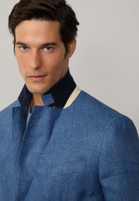 Giacca in denim blu con colletto alto, dotata di un accento blu scuro e bordo crema. Design semplice, tessuto texturizzato e dettagli di taglio visibili.
