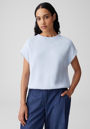 Frau mit langen dunklen Haaren trägt ein hellblaues, geripptes Kurzarmoberteil und dunkelblaue Hose, steht mit einer Hand in der Tasche vor einem grauen Hintergrund.