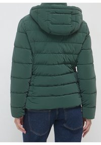 Giacca a vento verde scuro con cappuccio, caratterizzata da un design trapuntato e texture a costine. La vestibilità è corta con polsini elasticizzati e chiusura a zip.