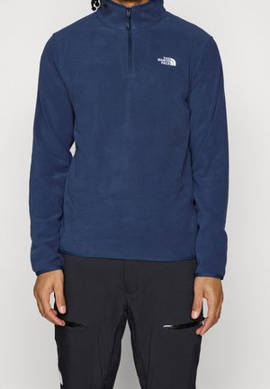 Osoba ubrana w granatowy polar z suwakiem do połowy wysokości z logo The North Face oraz czarne spodnie z kieszeniami na suwak.