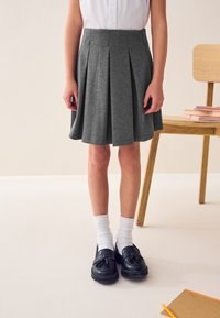 Jupe plissée grise en tissu doux, associée à des mocassins noirs et des chaussettes blanches. En arrière-plan, une chaise en bois et des livres.