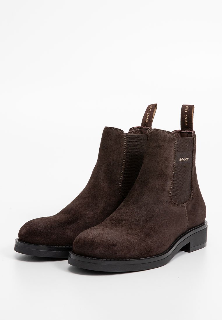 Botas de tobillo de ante marrón con paneles laterales elásticos, suela negra plana y lengüetas para ponerlas y quitarlas con facilidad. La textura parece suave y lisa.