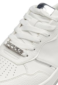 Zapatillas blancas con cuero texturizado y paneles lisos. Presenta cordones blancos, un acento metálico decorativo con gemas transparentes en la parte superior.