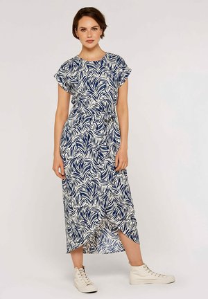 Maxikleid in Weiß und Marineblau mit einem Blättermuster, kurzen Ärmeln, einem Taillengürtel und einem seitlichen Schlitz. Kombiniert mit hellen, hochgeschnittenen Turnschuhen.