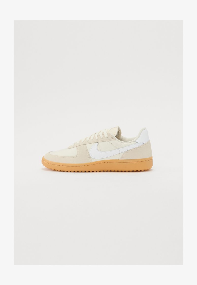 Beige textil felsőrészű sportcipők, fehér díszítéssel és gumitalpú, barna talppal. Jellemzője a kerek orr és a lapos fűző, amely biztos illeszkedést biztosít.