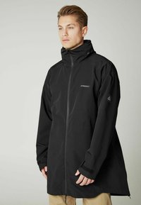 Čierny dlhý kabát s vysokým golierom, zipsom vpredu a nastaviteľnými manžetami. Obsahuje logo na hrudi a elegantný, minimalistický dizajn.