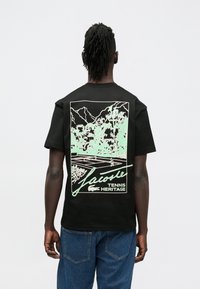 Juoda marškinėliai su mėtų žalumos grafika, vaizduojančia teniso sceną, kalnus ir apačioje užrašą „Lacoste Tennis Heritage“.