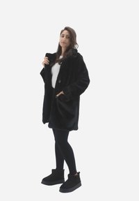 Sort faux fur frakke med en knap, sidelommer og en afslappet pasform, båret over en hvid top og parret med sorte leggings og ankelstøvler.