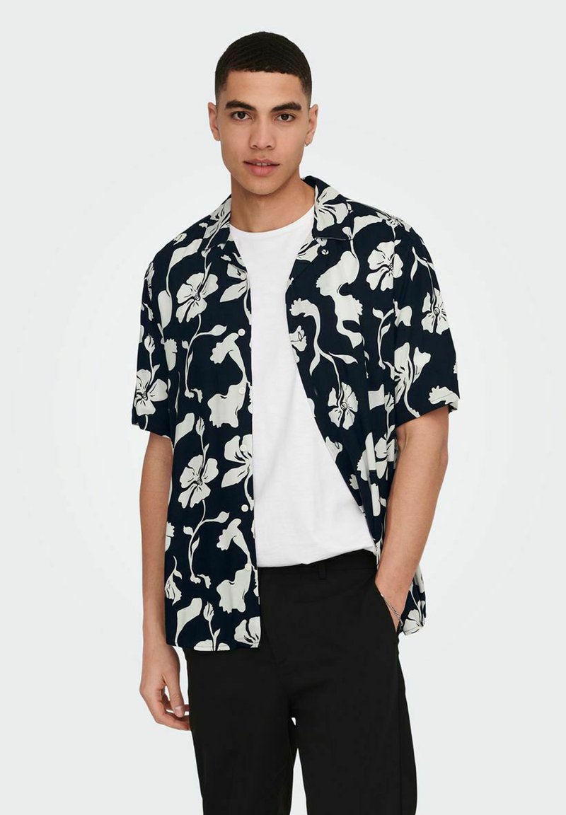 Kurzarmhemd in Marineblau mit weißem Blumenmuster, mit Knopfleiste und entspannter Passform, über einem weißen T-Shirt getragen. Schwarze Hose.