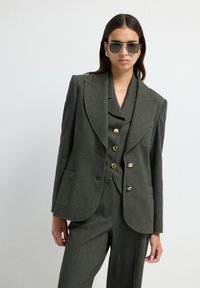 Olijfgroene op maat gemaakte blazer met scherpe revers, gouden knopen en zijzakken, gecombineerd met bijpassende broeken. Soepele textuur en gestructureerde pasvorm.