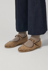 Zapatos Mary Jane de ante marrón con punta redonda, ribetes marrones y una correa con hebilla redonda, usados con calcetines metálicos dorados y pantalones de mezclilla oscuros.