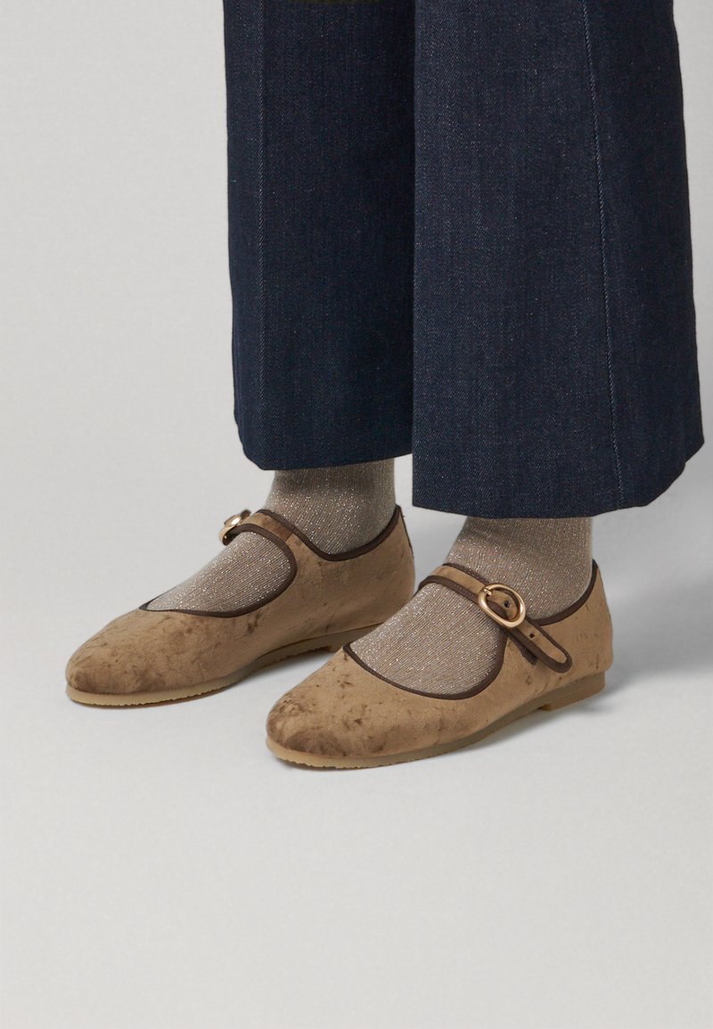 Zapatos Mary Jane de ante marrón con punta redonda, ribetes marrones y una correa con hebilla redonda, usados con calcetines metálicos dorados y pantalones de mezclilla oscuros.