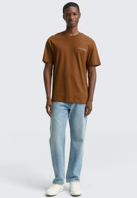 Bruine T-shirt met korte mouwen en ronde hals, met subtiele witte tekst. Gecombineerd met lichtblauwe rechte jeans en witte sneakers.