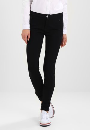 Person iført slanke sorte bukser, hvid skjorte med knapper og hvide sneakers med røde og blå stribede såler.