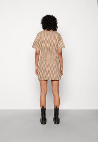 Vestido ajustado de mangas curtas em bege com um padrão castanho ondulado. Usado com botas pretas até o tornozelo. Fundo minimalista.