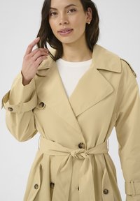 Trench coat bej cu un design cu două rânduri de nasturi, guler mare, curea legată și detalii cu nasturi. Fabricat dintr-un material neted, cu buzunare laterale.