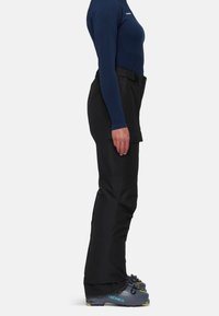 Pantalons de ski noirs avec une coupe droite, en tissu durable, poches latérales et une ceinture. Portés avec des bottes de ski grises présentant des accents jaunes.