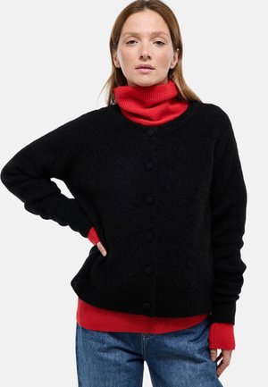 Cardigan noir en tissu duveteux avec boutons, porté sur un col roulé côtelé rouge. Comprend des manches longues et une coupe décontractée. Un jean en denim complète la tenue.