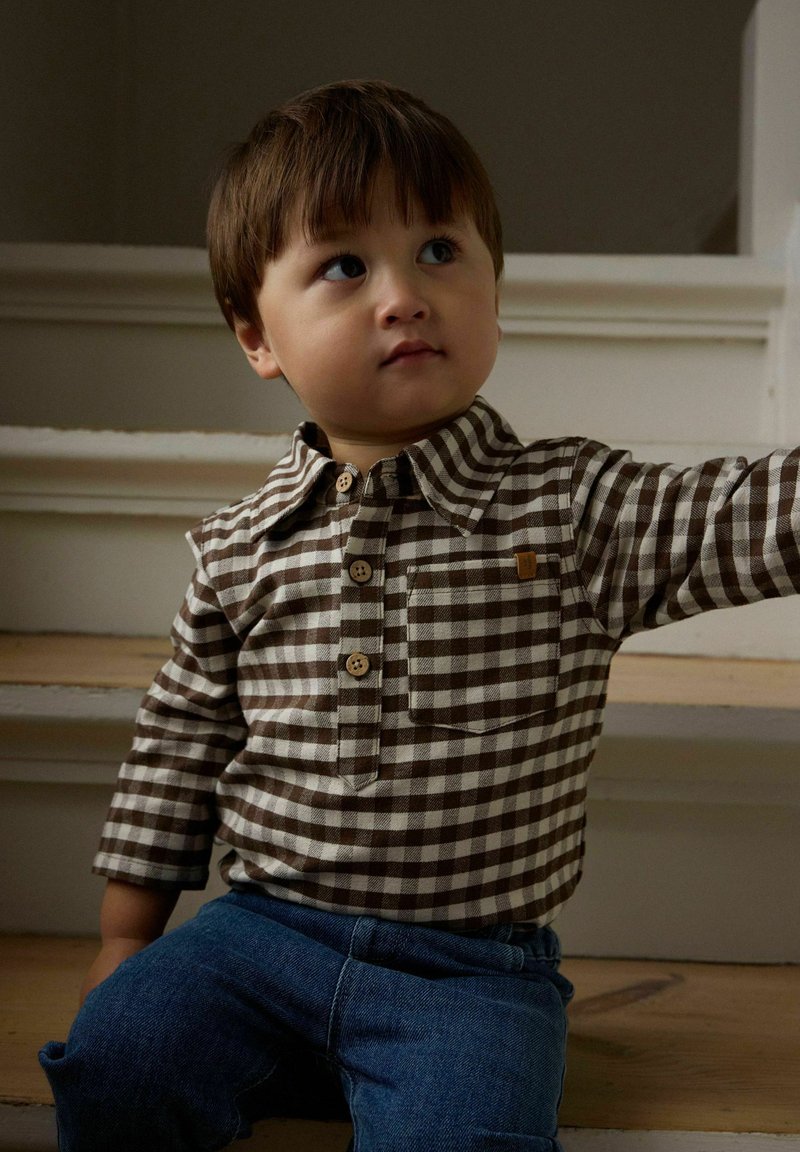 Jeune enfant aux cheveux courts et bruns portant une chemise à carreaux marron et blanche et un jean bleu, assis sur des escaliers en bois, regardant sur le côté.